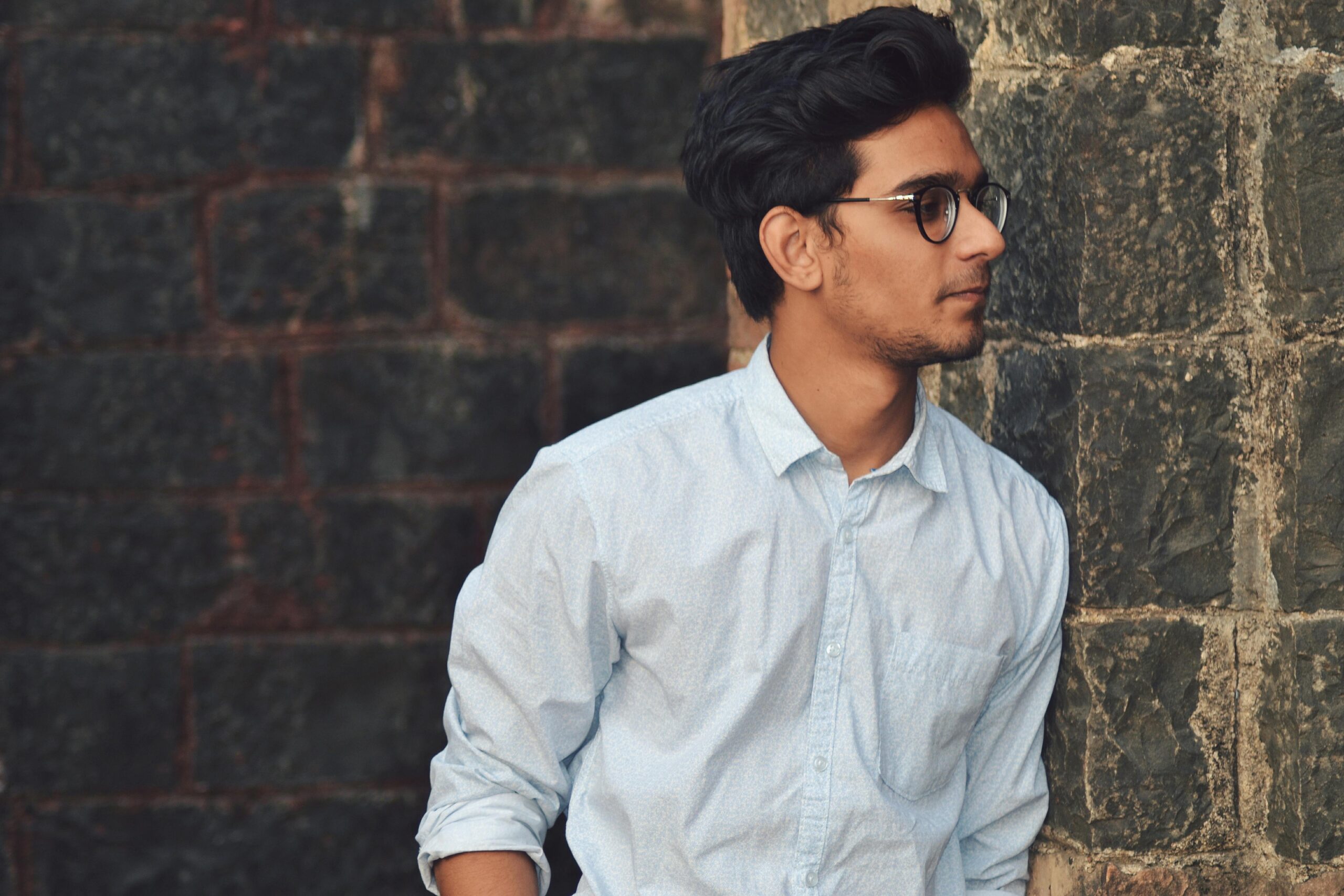 Digital Marketing Agency In Agra A stylish young man in glasses leans against a brick wall wearing a casual light blue shirt.